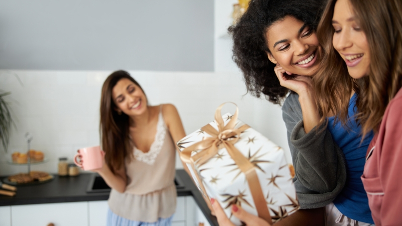 Three smiling friends share a wrapped present at home, showing the joy of thoughtful gifts for girls