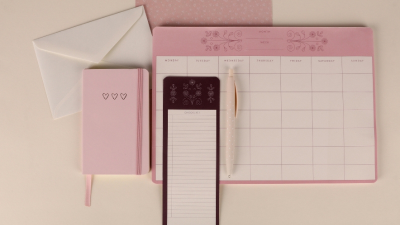 A pink stationery set with a notebook, planner, checklist pad, and pen is arranged on a desk