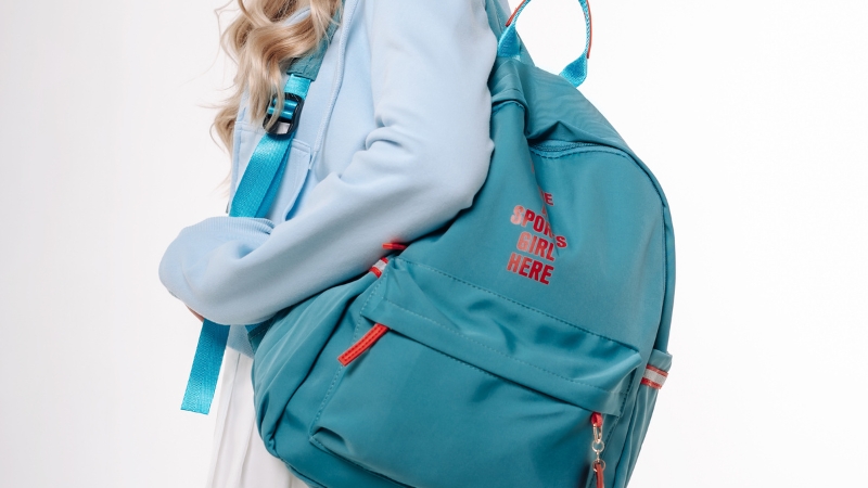 A girl carries a teal backpack with red zipper details and printed text on the side pocket
